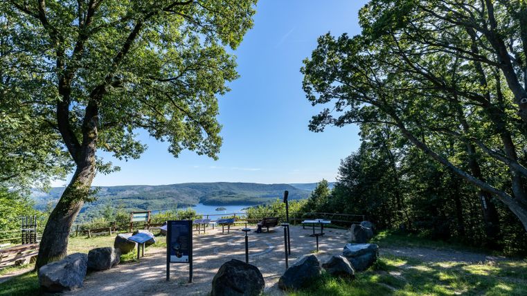 Een uitkijkpunt met uitzicht op een meer, omringd door bomen. De lucht is helder en het gebied is ingericht met stenen en paden.