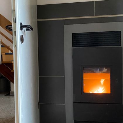 Ein moderner Kaminofen mit flammendem Feuer steht in der Küche. Eine offene Tür führt zu einer Treppe im Hintergrund.