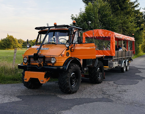 Een oranje Unimog met een aanhanger rijdt op een landelijke weg. In de aanhanger zitten personen en de omgeving is groen en bosrijk.
