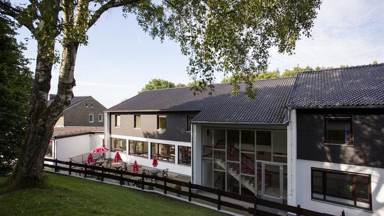 A modern building with a large window and an inviting entrance. Surrounded by trees and a green lawn, it offers a relaxed atmosphere.