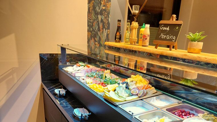 A buffet with a selection of fresh fruits, vegetables, and various side dishes. Bottles of sauces are provided along with a small board that reads "Good Morning."
