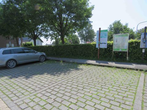 Ein Parkplatz mit einem geparkten silbernen Auto. Im Hintergrund sind Informationsschilder und eine grüne Hecke zu sehen.