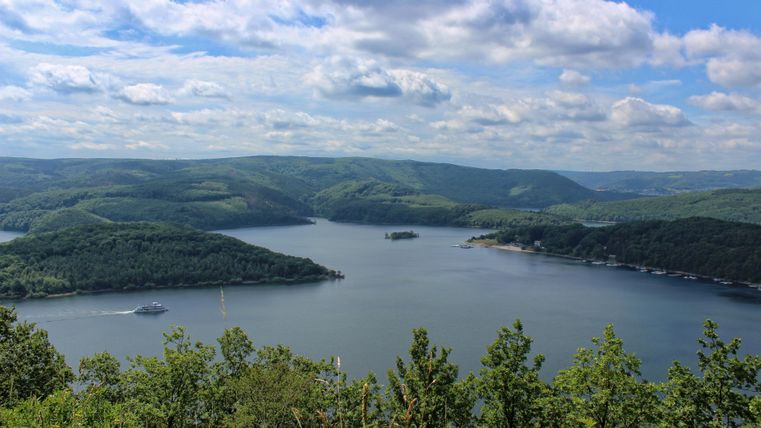 A picturesque landscape with a lake, surrounded by green hills. The sky is blue with some clouds.