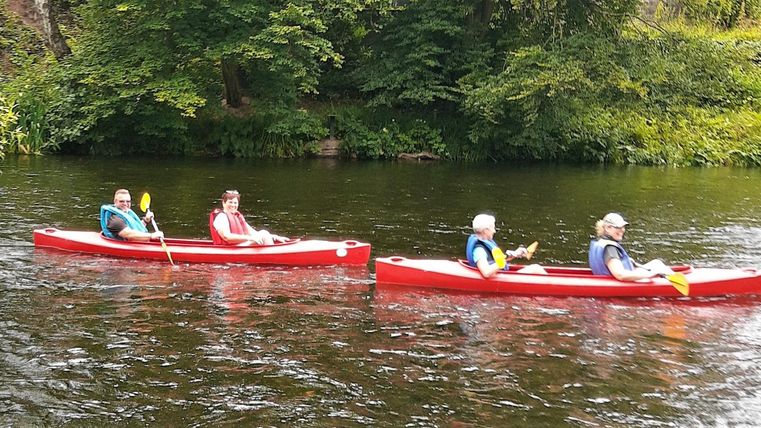 Vier Personen paddeln in roten Kanus auf einem ruhigen Fluss. Die Umgebung ist grün und üppig, umgeben von Bäumen.