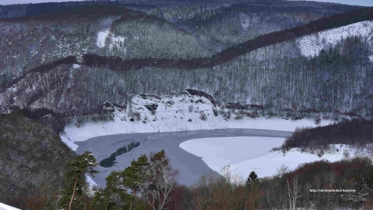 Een winterlandschap met een bevroren rivier en met sneeuw bedekte bomen. De heuvels op de achtergrond zijn begroeid met sparren.