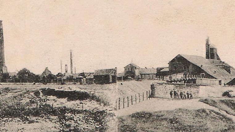 Een historische industrieaanzicht met fabrieksgebouwen en hoge schoorstenen. Op de voorgrond is een oneffen landschap met hekken en enkele mensen te zien.