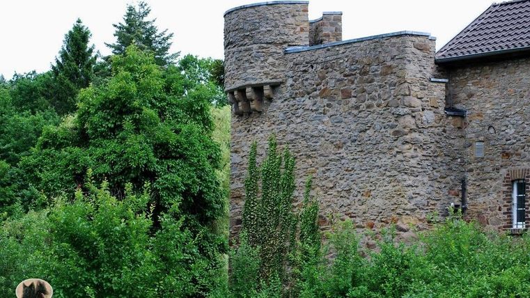 Een oude stenen structuur met een ronde toren, omringd door weelderig groen. Het landschap is rustig en natuurlijk.