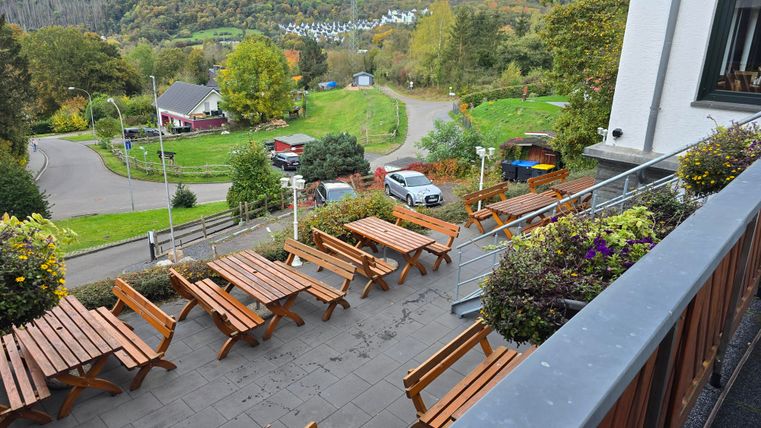A cozy terrace with wooden tables and benches. In the background, a green landscape stretches out with lush trees and gentle hills.