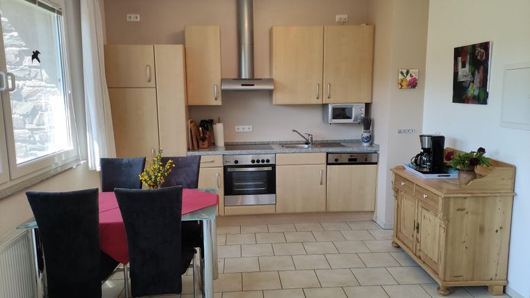 A bright, modern kitchen with wooden cabinets and a dining table for four people. In the background, kitchen appliances and a window front can be seen.