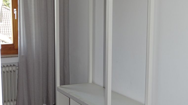 Een eenvoudige, witte kledingstandaard met houten kledinghangers staat tegen de muur. Daaronder bevindt zich een plank met open vakken.