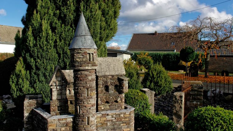 Een prachtig stenen kasteel met een toren in een tuin. Op de achtergrond zijn groene bomen en een huis zichtbaar.