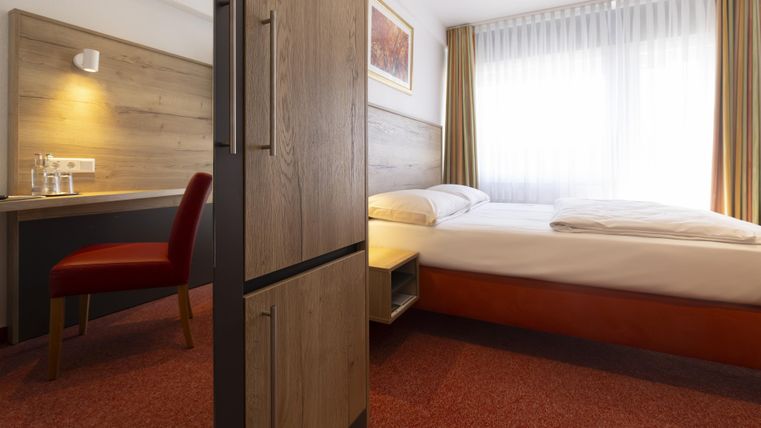 Ein modernes Hotelzimmer mit einem Doppelbett und einem Schreibtisch. Das Zimmer hat große Fenster und eine freundliche Einrichtung.