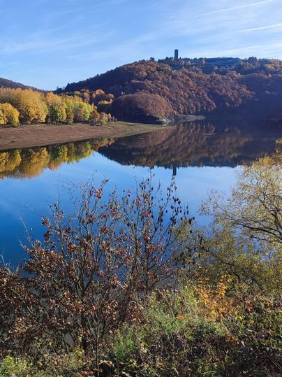 A tranquil lake with clear reflection and colorful autumnal shore. In the background, a hill rises with a lookout tower.