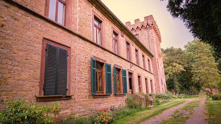 Een historisch gebouw met stenen gevel en groene luiken. Het pad leidt door een tuin met weelderige vegetatie.