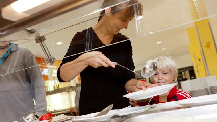 A woman serves food at a table in a cafeteria. A happy child smiles up at her.