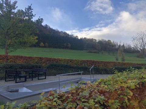 Eine ruhige Landschaft mit einer Aussicht auf grüne Wiesen und Bäume. Im Vordergrund stehen moderne Sitzgelegenheiten auf einem gepflasterten Bereich.