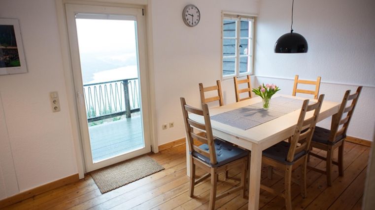 A bright dining room with a white table and wooden chairs. A flower in the center of the table and access to a balcony with a view.