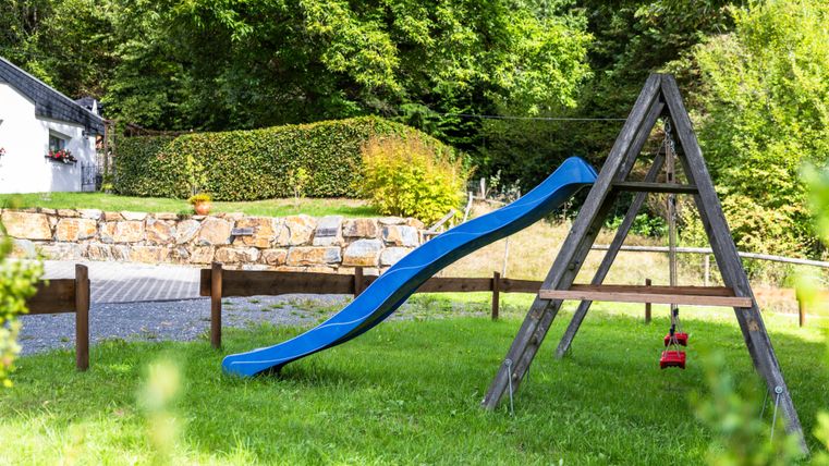Een glijbaan in een groene tuin met bomen op de achtergrond. De speeltuin is rustig en uitnodigend.