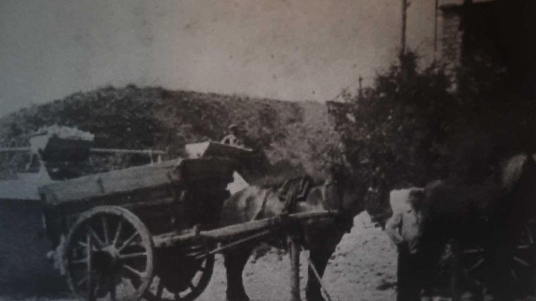 Een paardenkar staat stil op een weg. De wagen is met verschillende goederen beladen en wordt door paarden getrokken.