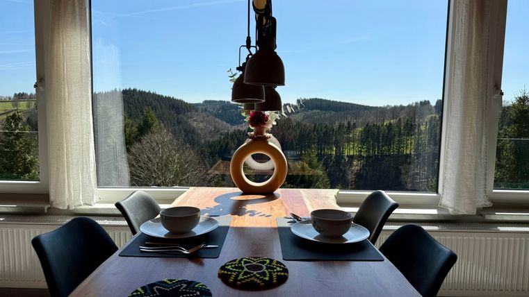 Een lichte eetruimte met een tafel en twee schalen. Buiten zie je een pittoresk landschap met heuvels en een heldere blauwe lucht.