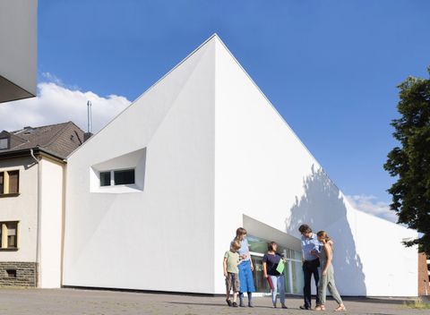 Ein modernes, weißes Gebäude mit einer markanten, dreieckigen Form. Vor dem Gebäude stehen mehrere Personen, die miteinander interagieren.