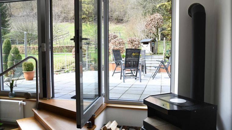 A cozy room with large windows overlooking an outdoor terrace. Outside, there are seating furniture and a green landscape visible.