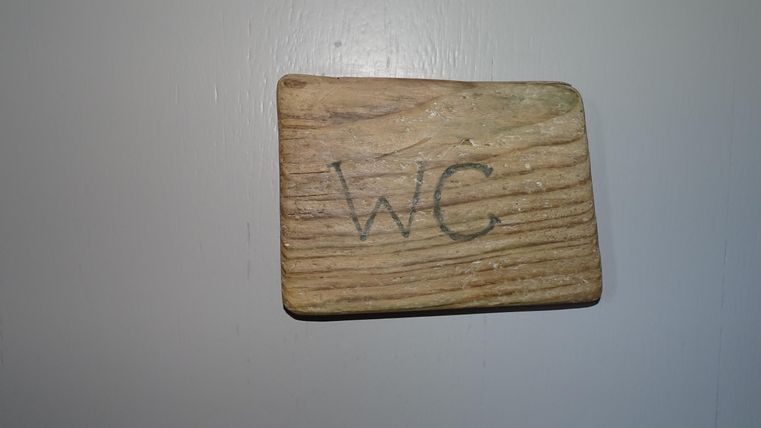 Een houten bord met de tekst "WC". Het is aan een muur bevestigd.