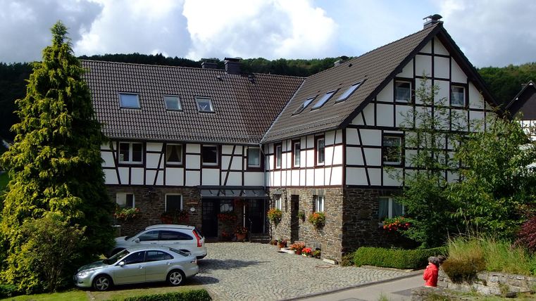 A charming half-timbered building with a large garden. In front of the house, there are a few cars and there are green trees in the background.