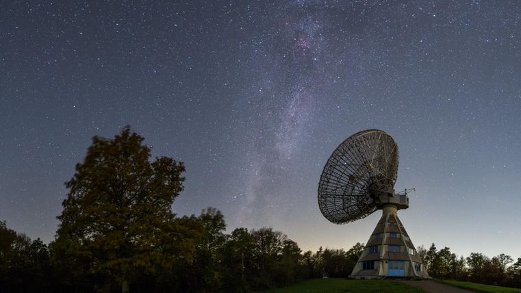 Een grote radiotelescoop staat onder een heldere nachtelijke hemel met de Melkweg. Bomen omringen de installatie in een rustige landschap.