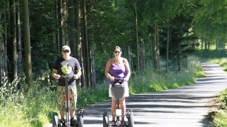 Two people are riding on Segways along a forest path. The surroundings are green and sunny.
