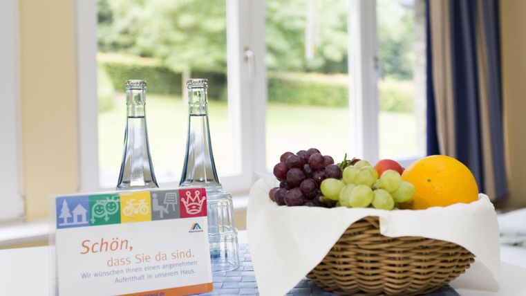 A table with two bottles of water and a fruit bowl containing grapes, an orange, and apples. In the background are large windows overlooking a green garden.