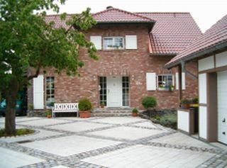 A pretty two-story house made of red bricks. In front of the house, there is a paved entrance area with a bench and trees.