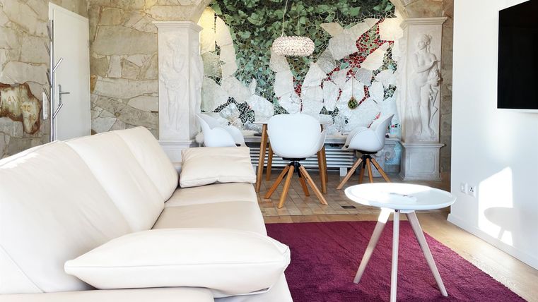 A modern living room with a white sofa and a purple rug. In the background, there is a beautiful dining area with an artistic mural.