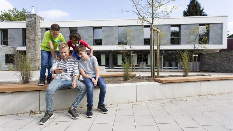 Four children are sitting and standing in a modern environment. They are looking together at a mobile phone and appear to be interested and entertained.