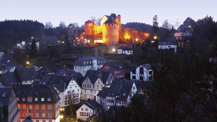 Een pittoreske stad met historische gebouwen onder een heldere lucht. Op de achtergrond straalt een kasteel in warm licht.