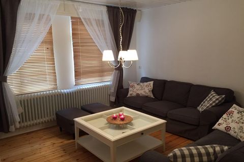 A cozy living room with a gray sofa and a white table. Natural lighting from windows with blinds gives the room a bright atmosphere.