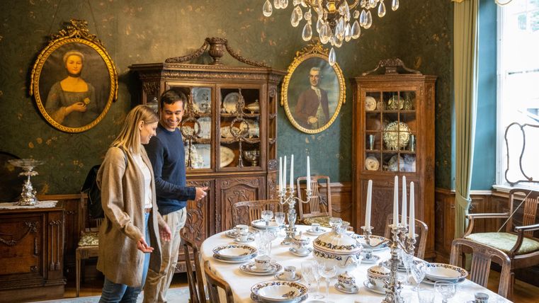 A stylishly furnished dining room with an elegant table set with fine china. In the background, antique furniture and portraits on the wall can be seen.