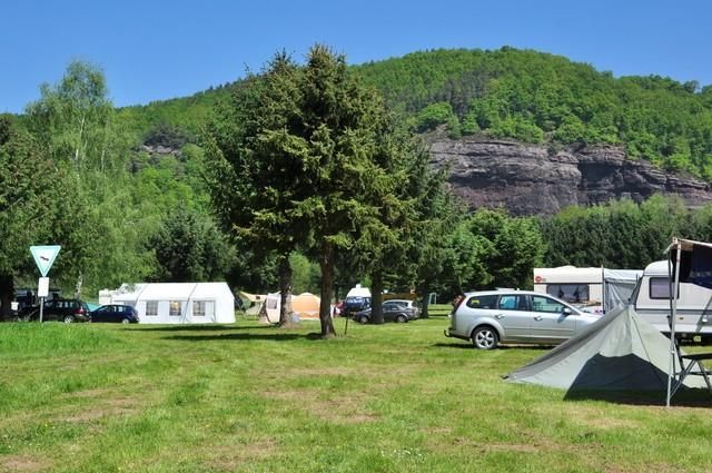 Een camping met groene weiden, bomen en caravans. Op de achtergrond rijst een bosrijke berglandschap op onder een blauwe lucht.