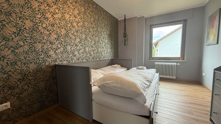 Een moderne slaapkamer met een groot tweepersoonsbed en een aantrekkelijk plantenpatroon op de muur. Het heeft een raam dat daglicht binnenlaat en een houten vloer.