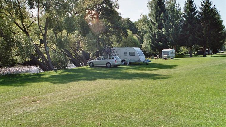 Een rustige camping aan het water met groene weiden. Twee campers staan onder schaduwrijke bomen.