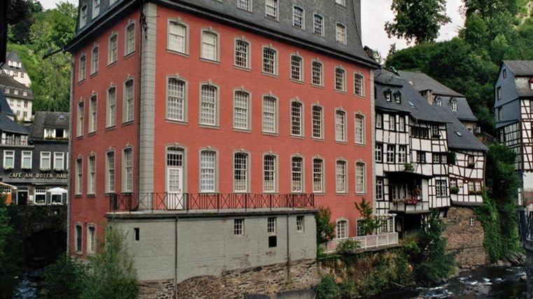 A historic building with a red facade next to a small river. In the background, more picturesque houses can be seen, surrounded by green trees.
