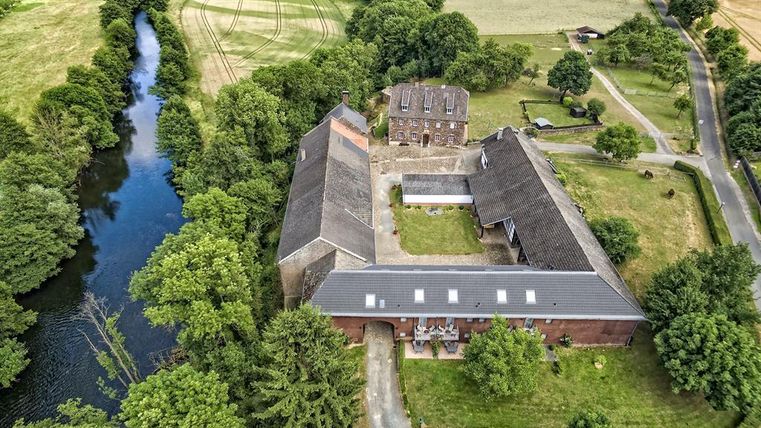 Eine Luftaufnahme eines rustikalen Gebäudes entlang eines Flusses, umgeben von üppigem Grün. Im Hintergrund sind Felder und Bäume zu sehen.