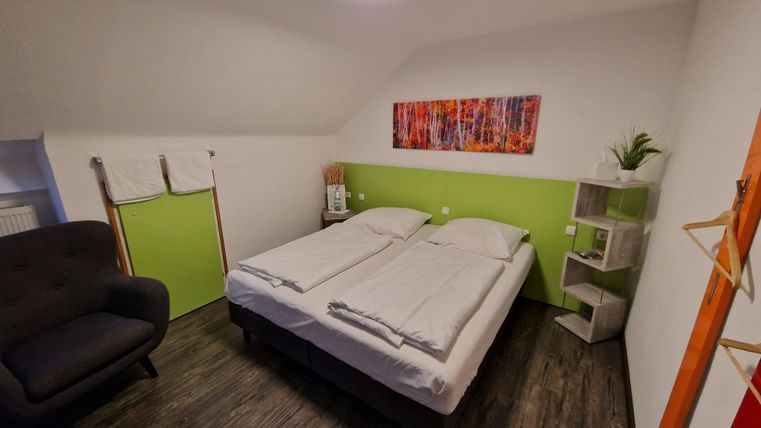 A cozy bedroom with a double bed and a green wall. A seating area with an armchair and a modern shelf is also available.