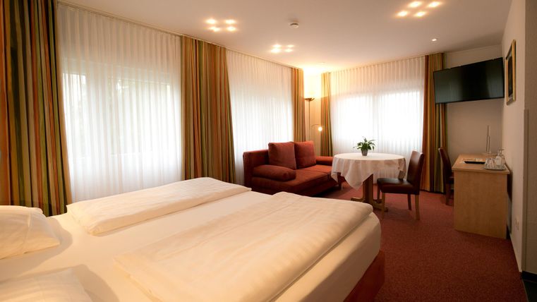 A modern hotel room with two beds, a sofa, and a table. The room has large windows with curtains and a television on the wall.