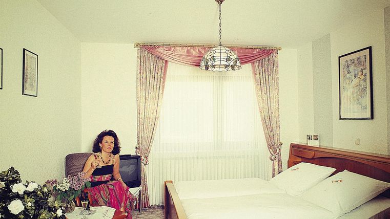 Een gezellige slaapkamer met een tweepersoonsbed en een fauteuil. Een vrouw zit ontspannen in de ruimte naast een bloemenarrangement.