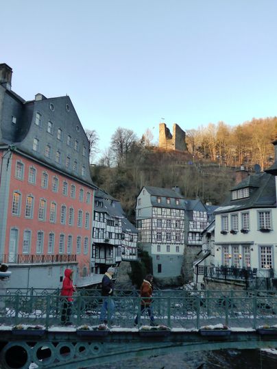 A picturesque old town with historic half-timbered houses and a bridge over a calm river. In the background, a castle can be seen on a hill.