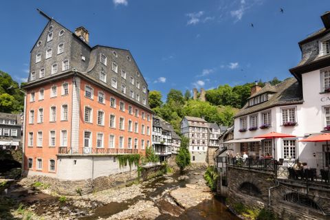 A picturesque view of the town with historical buildings along a small river. In the background, wooded hills and a blue sky can be seen.