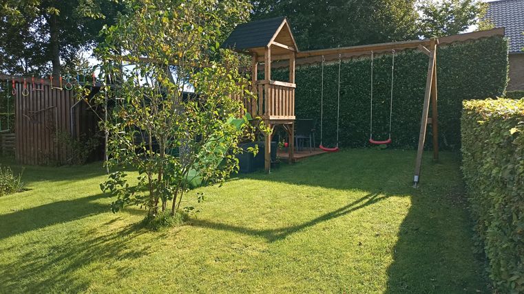 Ein schöner Garten mit einer Spielturmanlage und einer Schaukel. Die grüne Wiese ist von Hecken umgeben und es scheint sonnig zu sein.