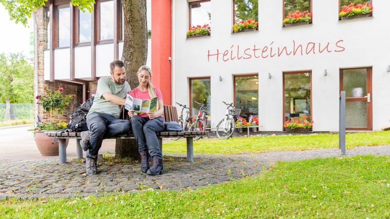 Zwei Personen sitzen auf einer Bank unter einem Baum und lesen gemeinsam eine Karte. Im Hintergrund ist das Heilsteinhaus mit blühenden Fenstern und Fahrrädern zu sehen.