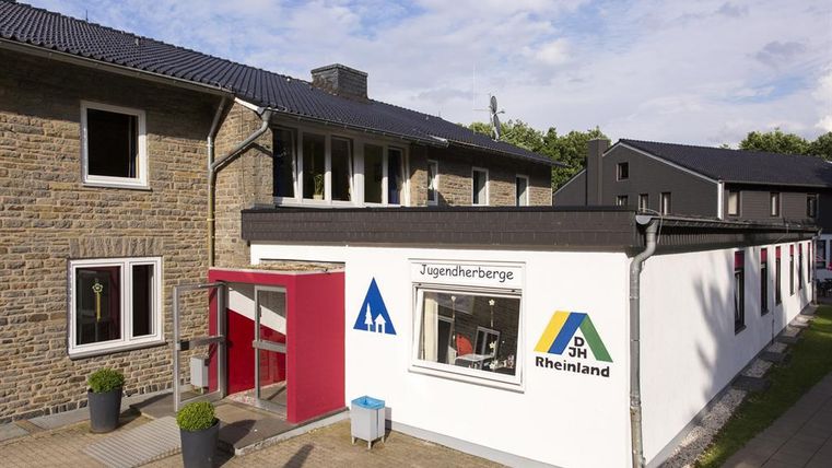 A modern youth center with two buildings and a welcoming entrance area. The façade is partly white and displays the logo of the Youth Hostels Rheinland.